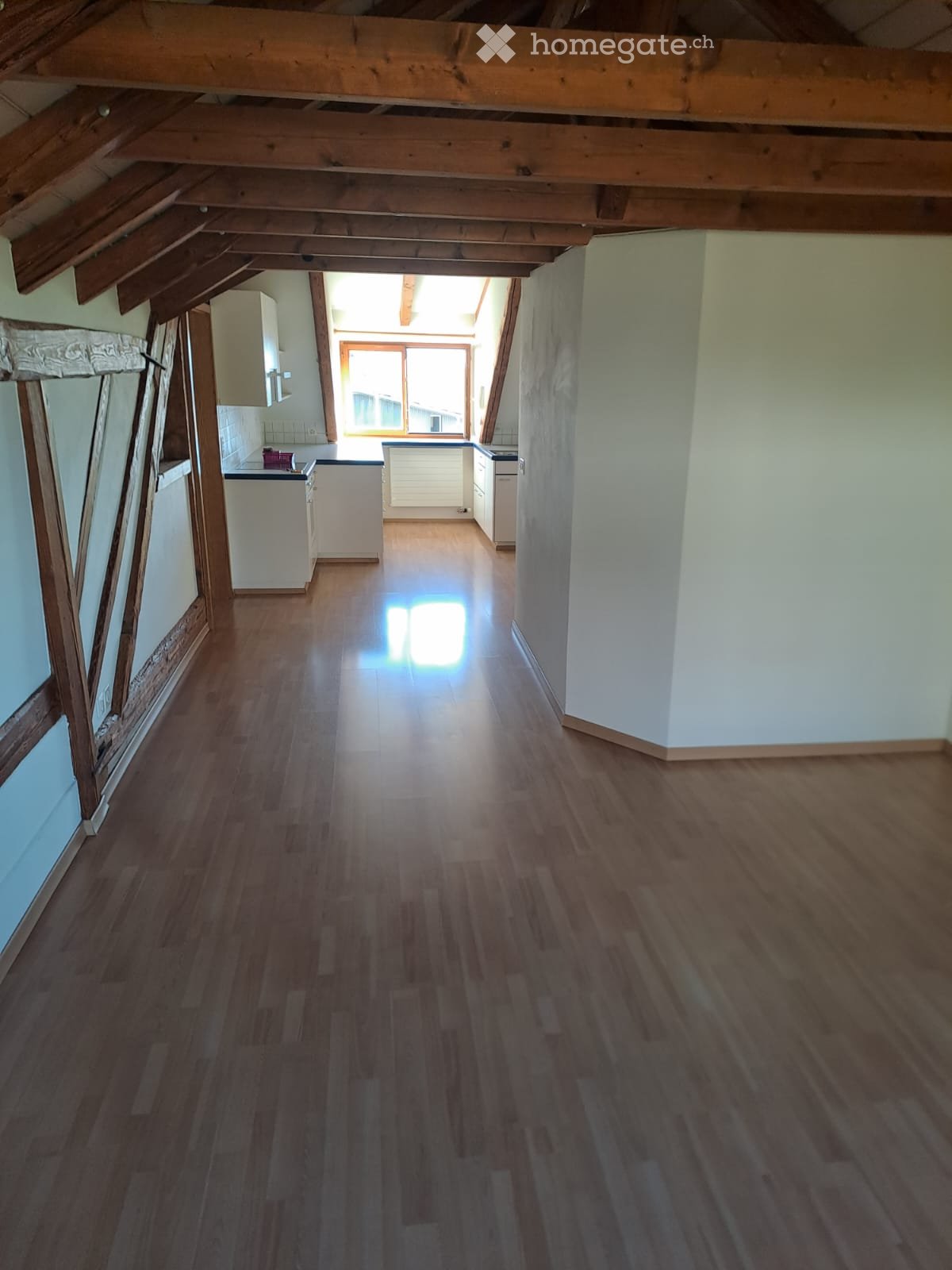 Empty hallway with wooden flooring, white walls, and exposed wooden beams on the ceiling. Natural light streams in from a window at the end of the corridor, illuminating the space.