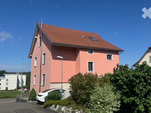 Two-story house with pink exterior, red tile roof, white-framed windows, and a small garden area with a parked car.
