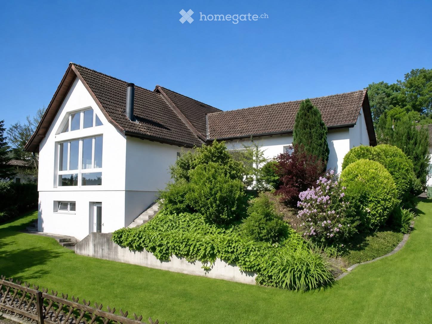Two-story white house with a brown tiled roof, large windows, and a well-maintained garden with green lawn and various shrubs and trees.