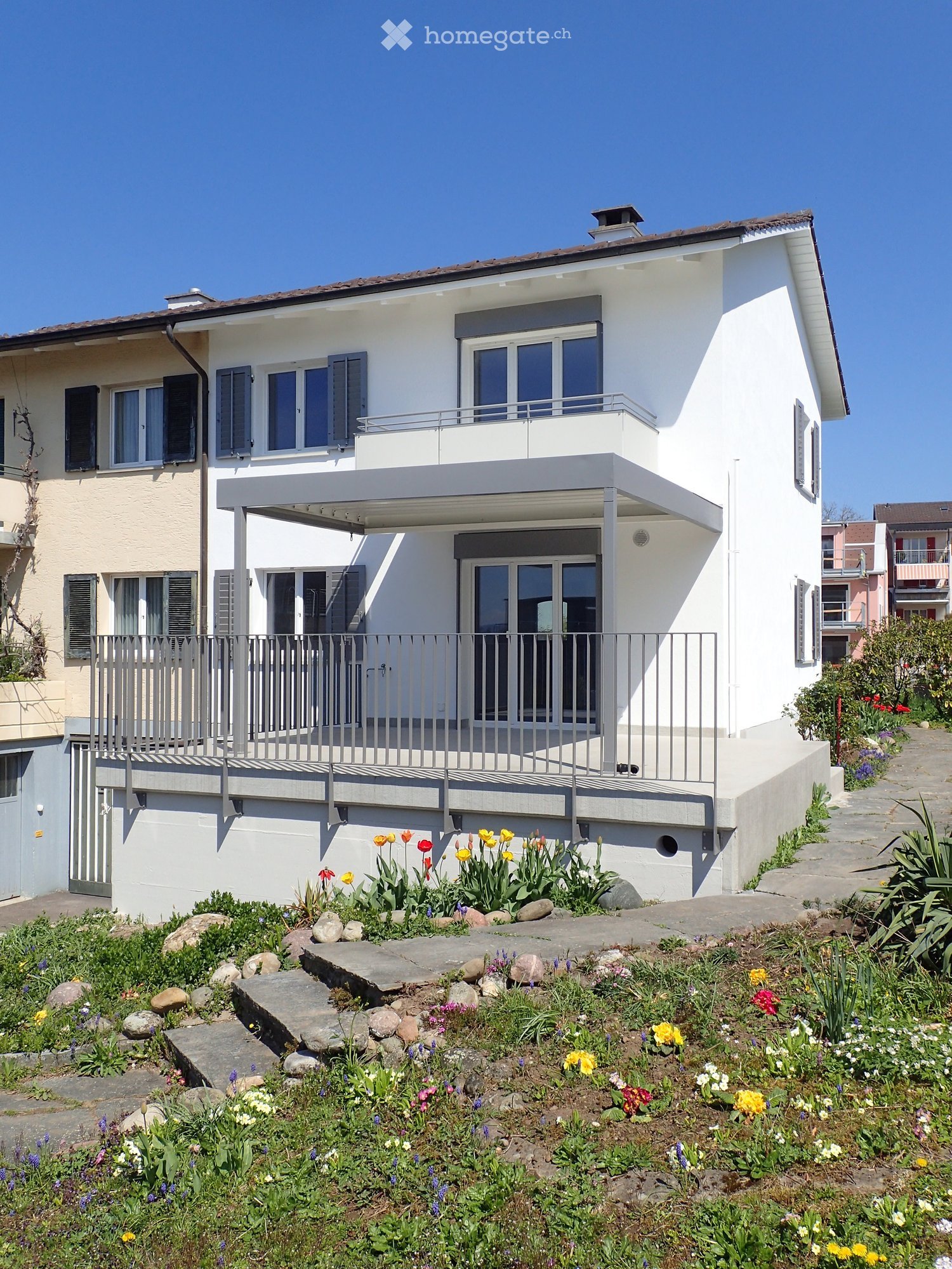 Modern two-story house with white exterior, large windows, and a balcony. The front yard features a garden with colorful flowers and stepping stones.