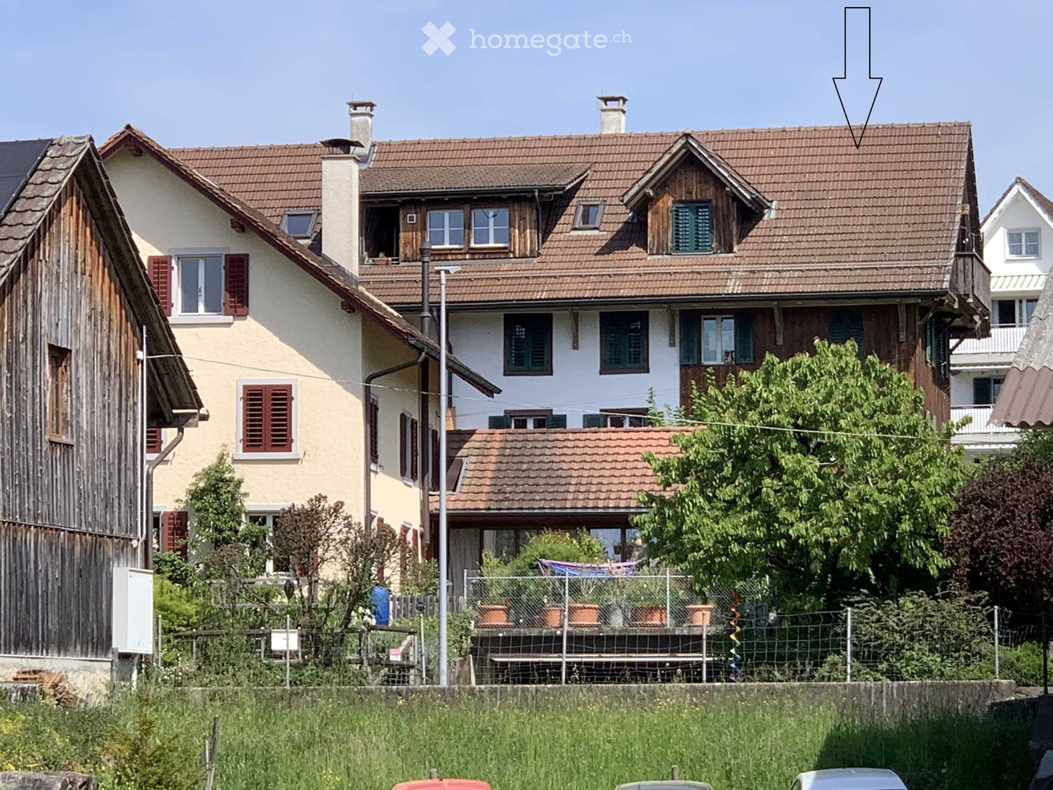 A two-story house with a tiled roof, multiple chimneys, and several windows with shutters. The house has a mix of white and wooden exterior walls. There is a fenced garden area with potted plants in front of the house.