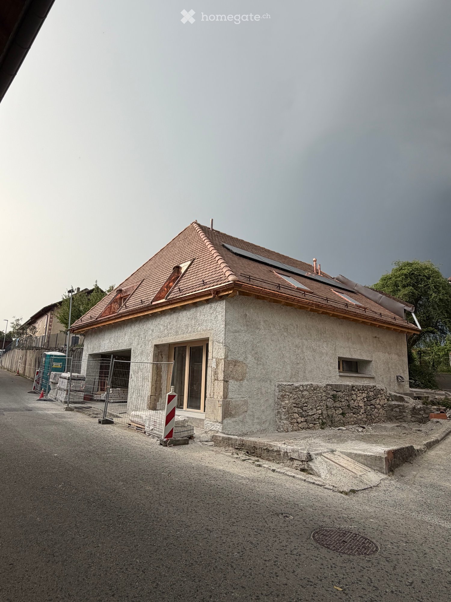 A small, single-story building with a tiled roof and concrete walls, featuring a fenced-off area and construction materials nearby.