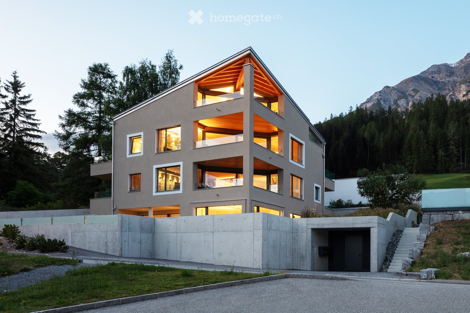 Modern, multi-story house with large windows and a raised ground floor, surrounded by trees and mountains.