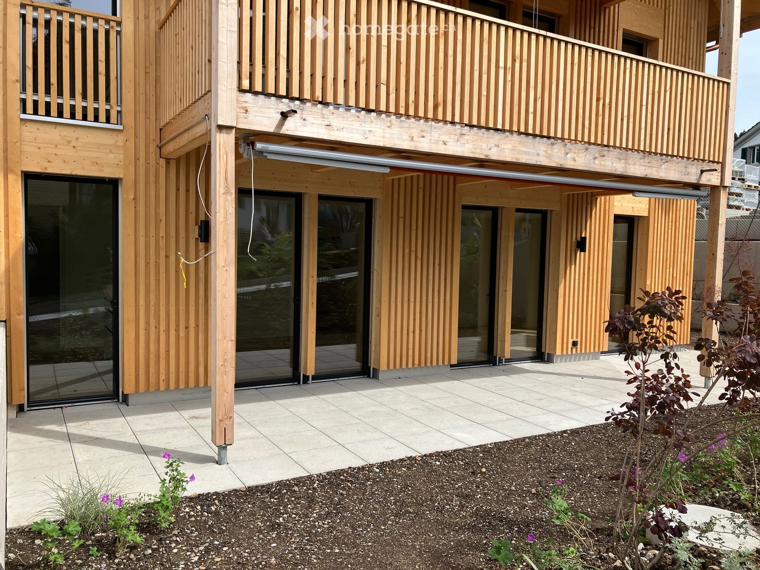 Modern wooden building with large glass doors and balconies, surrounded by small garden area with plants and flowers.