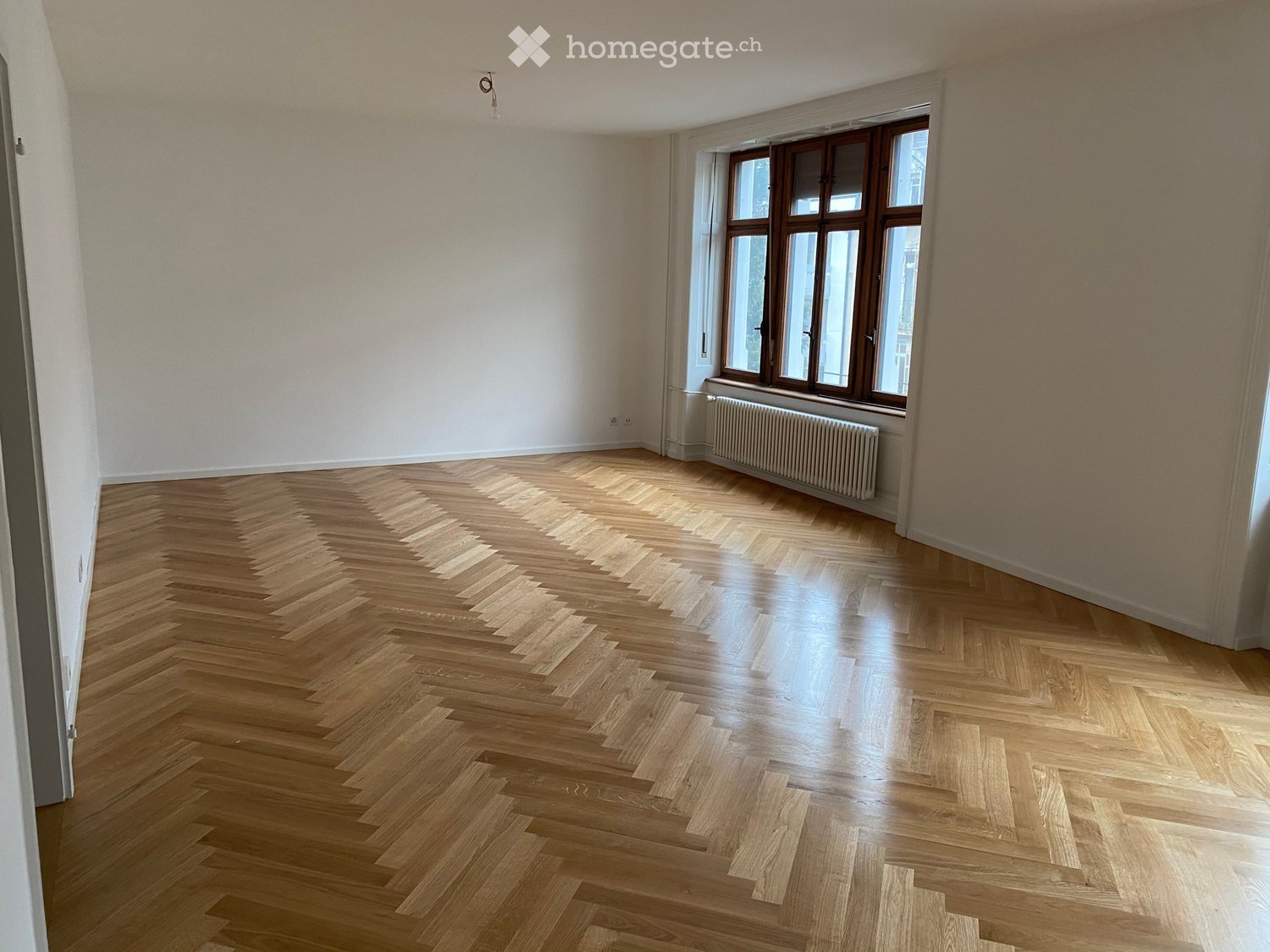 Empty room with herringbone-patterned wooden flooring, large windows with wooden frames, and a radiator beneath the windows.