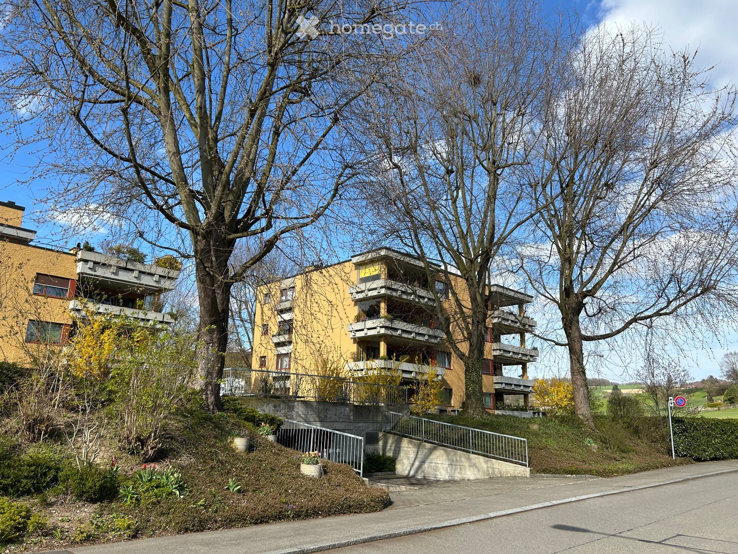 Multi-story yellow apartment building with balconies, surrounded by trees and greenery, with a raised ground floor entrance.