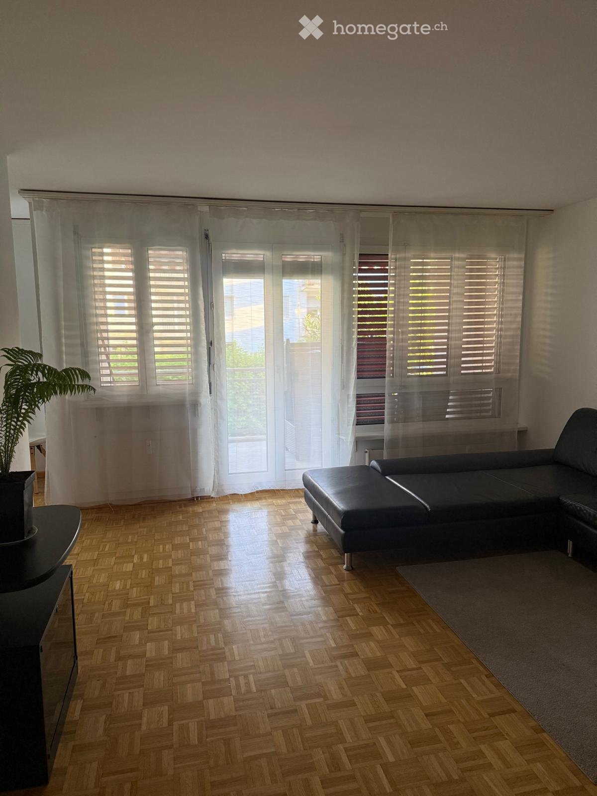 Living room with wooden herringbone flooring, large windows with blinds, a black sectional sofa, a small black table, a potted plant, and a grey rug.