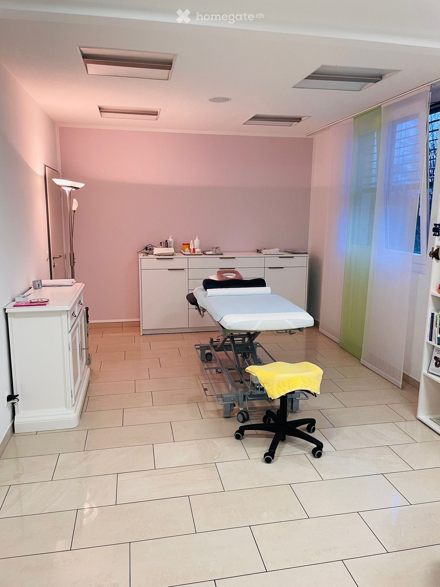 A clean, well-lit room with a treatment table, a stool, cabinets, a lamp, and a mirror. The walls are light pink, and the floor is tiled.