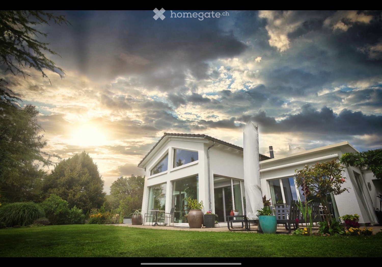white two-story house, large windows, outdoor seating area with chairs and table, potted plants, garden with lush green lawn, trees and bushes, cloudy sky in the background