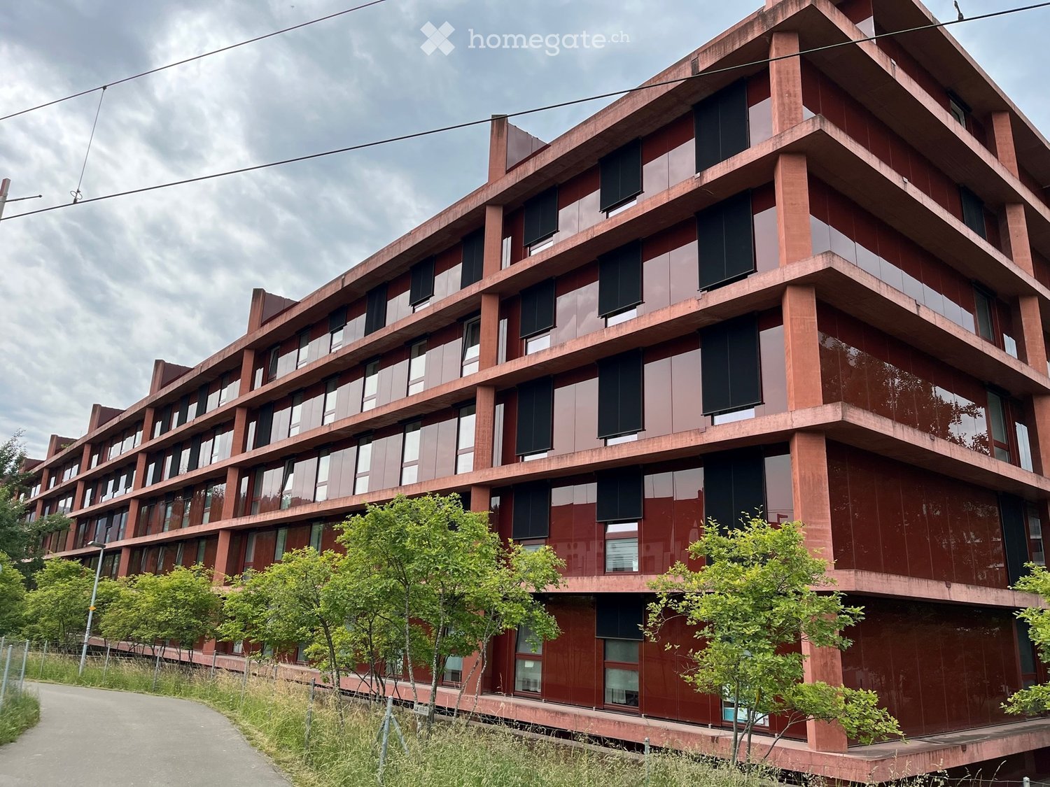 Modern multi-story building with red brick facade, large windows, and balconies. Surrounded by greenery and a paved pathway.