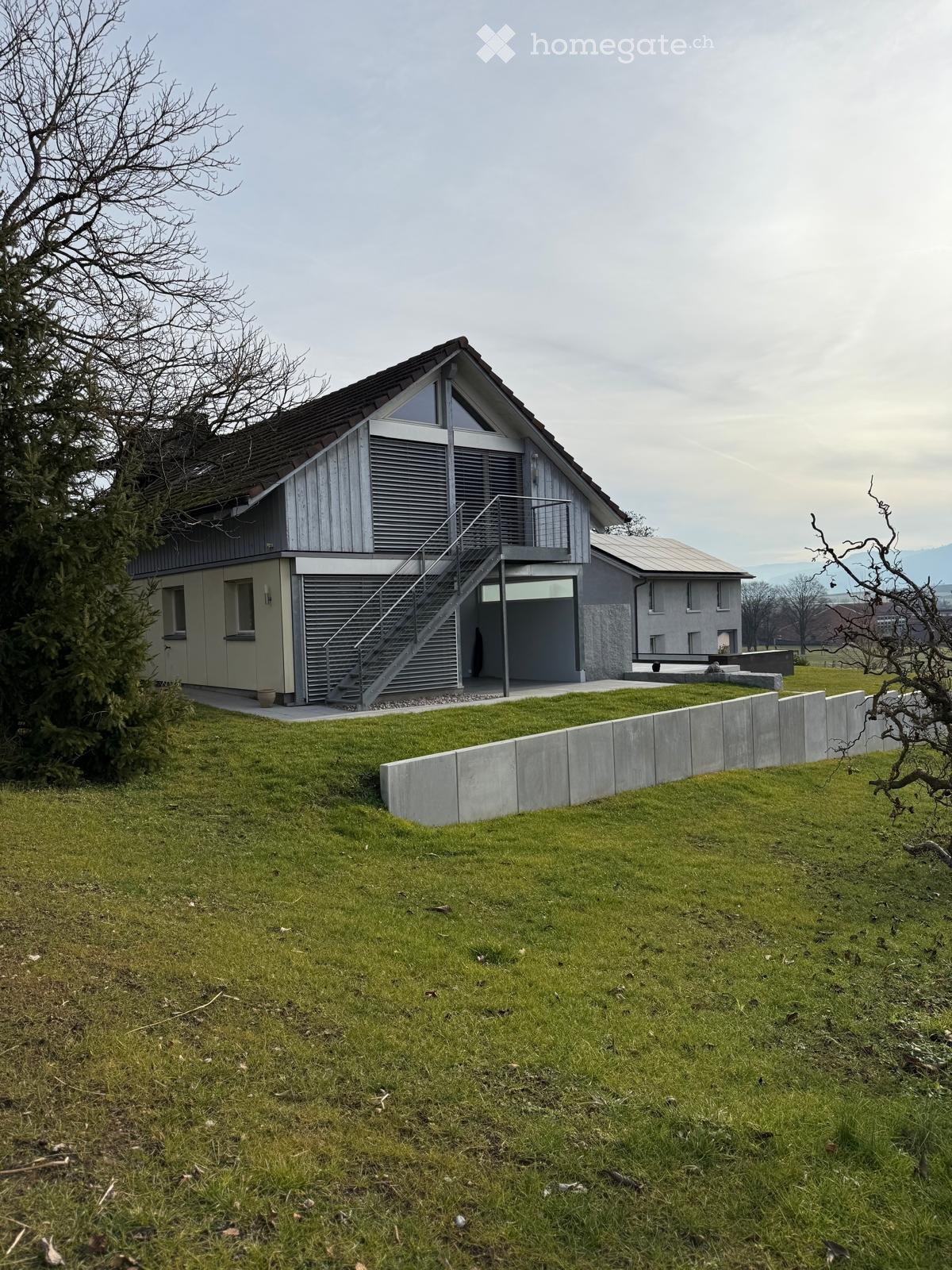 Modern 2 story house, grey and white facade, large windows, gray roof, driveway, large grass field, concrete barrier