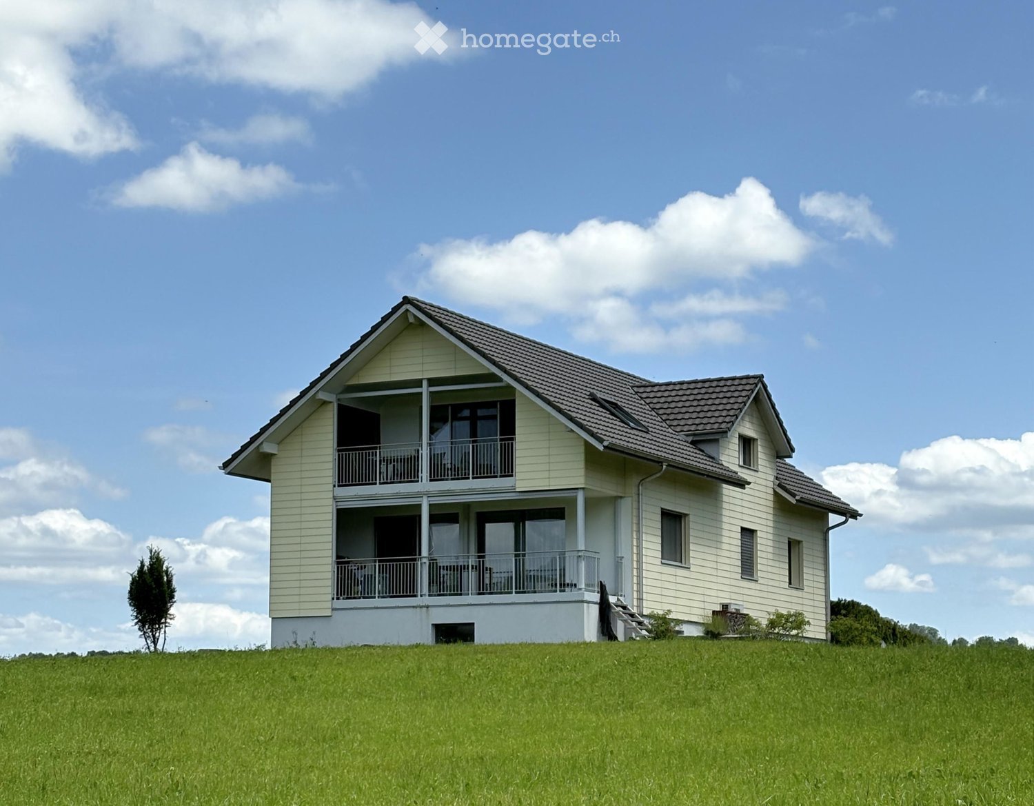 Two-story house with light yellow siding, dark brown roof, multiple windows, and balconies on the upper level.