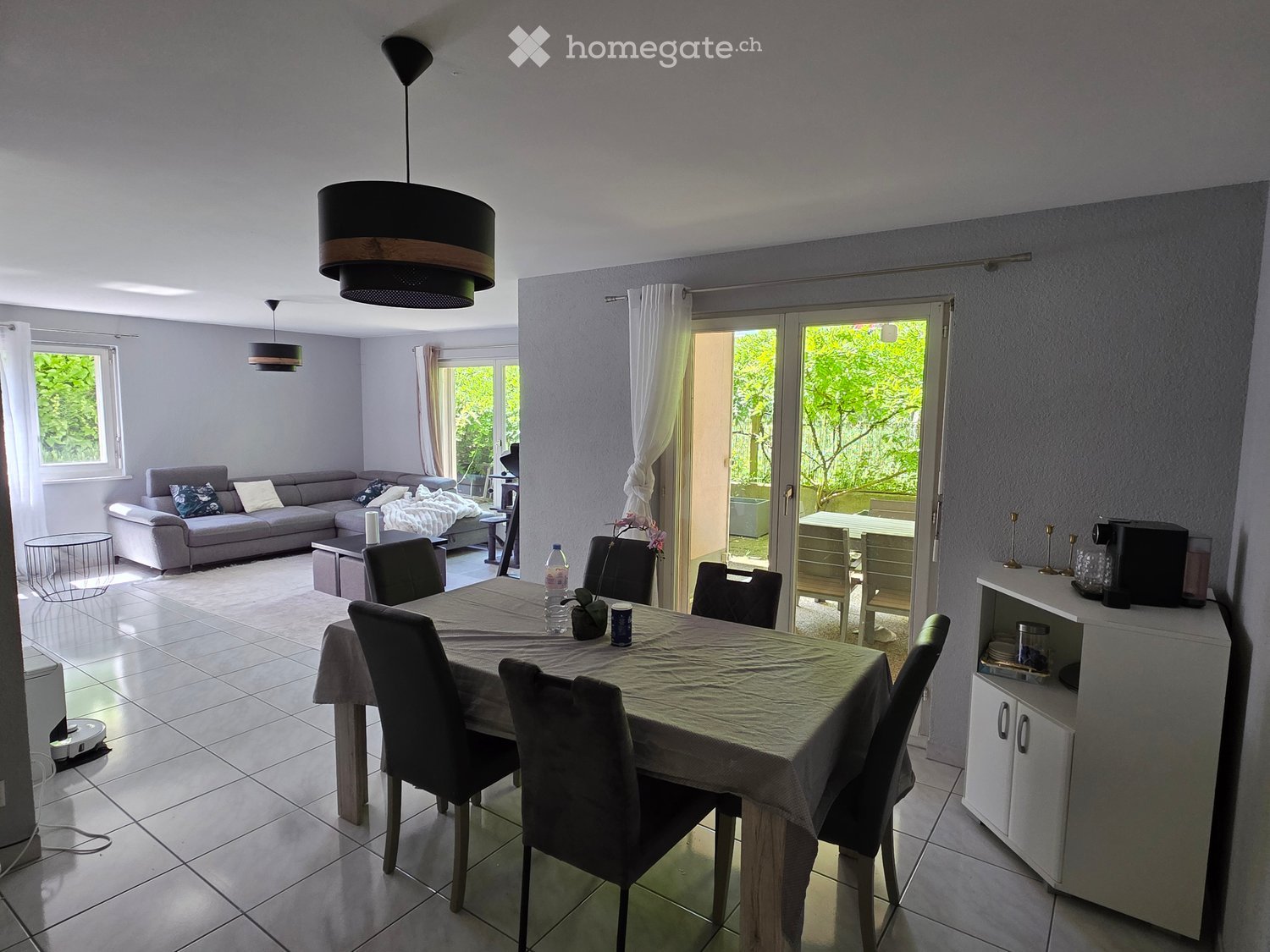 Bright living and dining area with a grey sofa, coffee table, dining table with chairs, and a small cabinet with a coffee machine. Large windows and sliding glass doors lead to a balcony.
