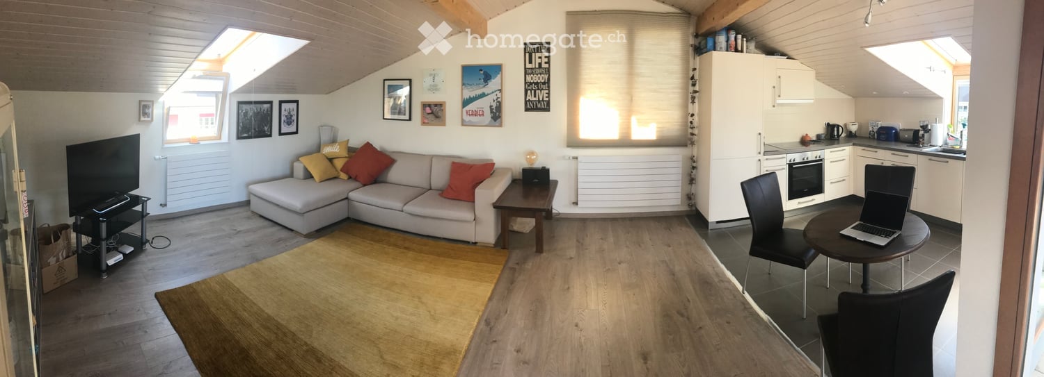 Bright, open-plan living and dining area with wooden flooring, a grey sectional sofa with colorful cushions, a coffee table, a TV stand, a small dining table with chairs, and a compact kitchen area with white cabinets, a fridge, oven, microwave, and a cou
