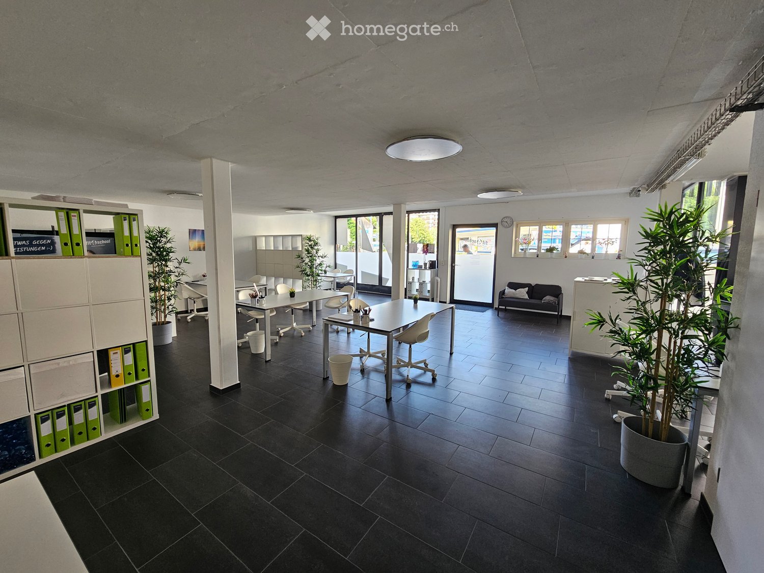Modern office space with black tiled flooring, white walls, and large windows. Features include desks, chairs, a sofa, potted plants, and storage cabinets.