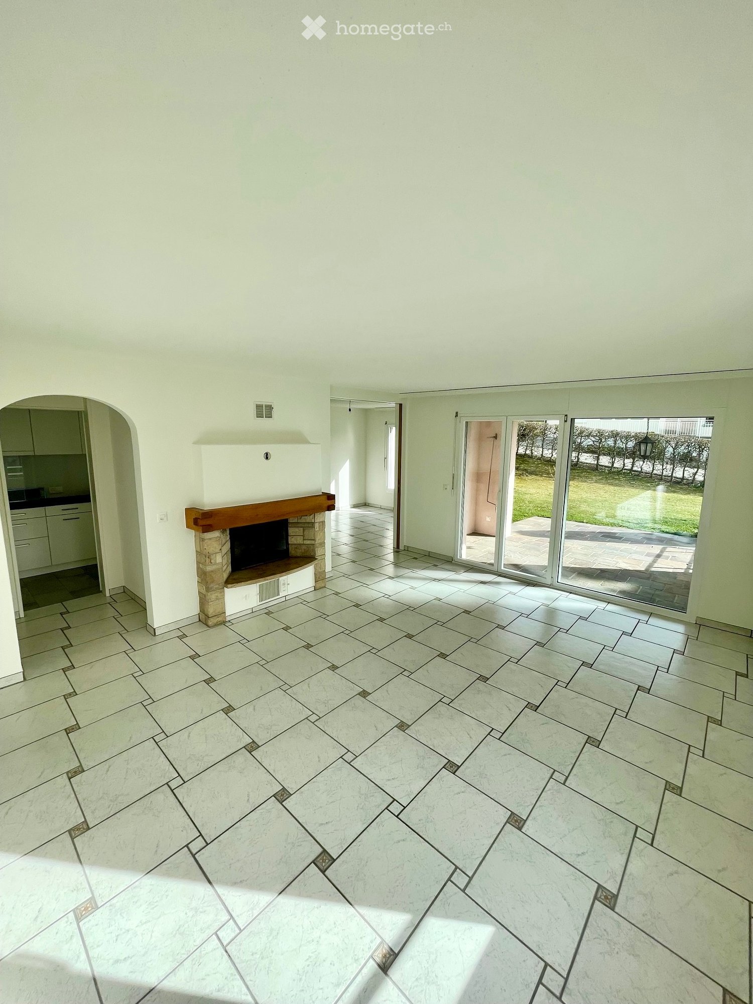 Empty living room with tiled floor, fireplace, sliding glass doors leading to outdoor area, and an adjacent kitchen area visible through an archway.