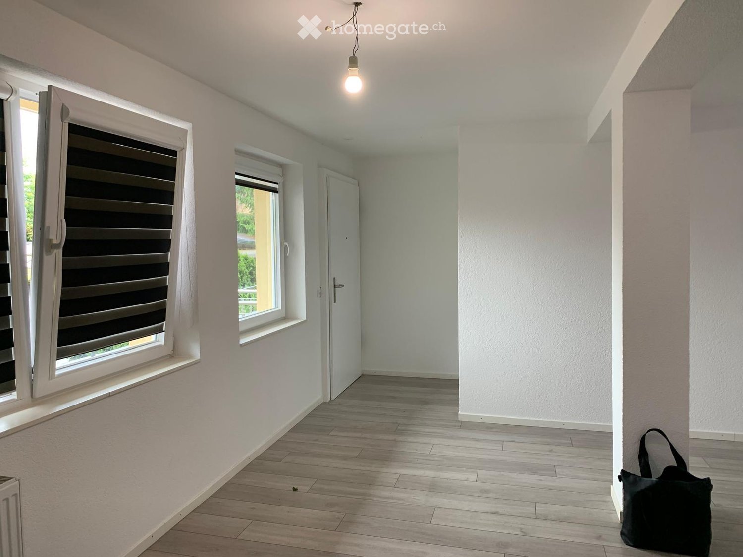 Empty hallway with light-colored walls, wooden flooring, and white-framed windows with blinds. A single black bag is placed near the wall.