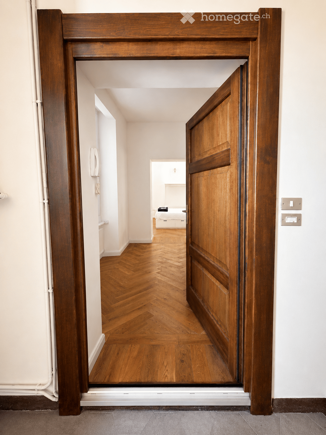 A hallway with wooden doors and herringbone-patterned wooden flooring, leading to a bedroom with a bed and nightstand visible in the background.