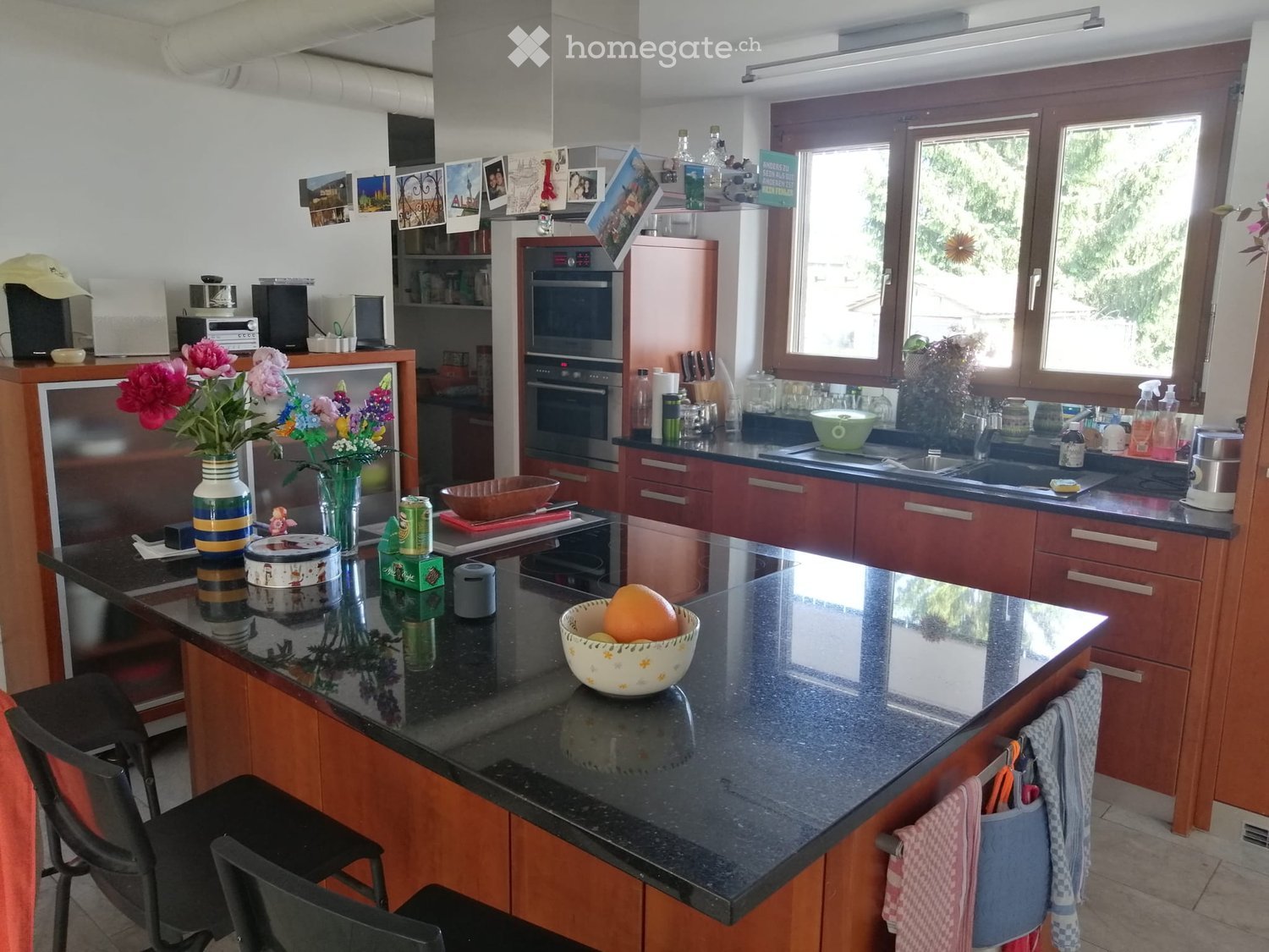 Modern kitchen with wooden cabinets, granite countertop, and various appliances including a refrigerator, oven, and microwave. There is a bowl of fruit on the island, a vase with flowers, and a sink with a faucet.