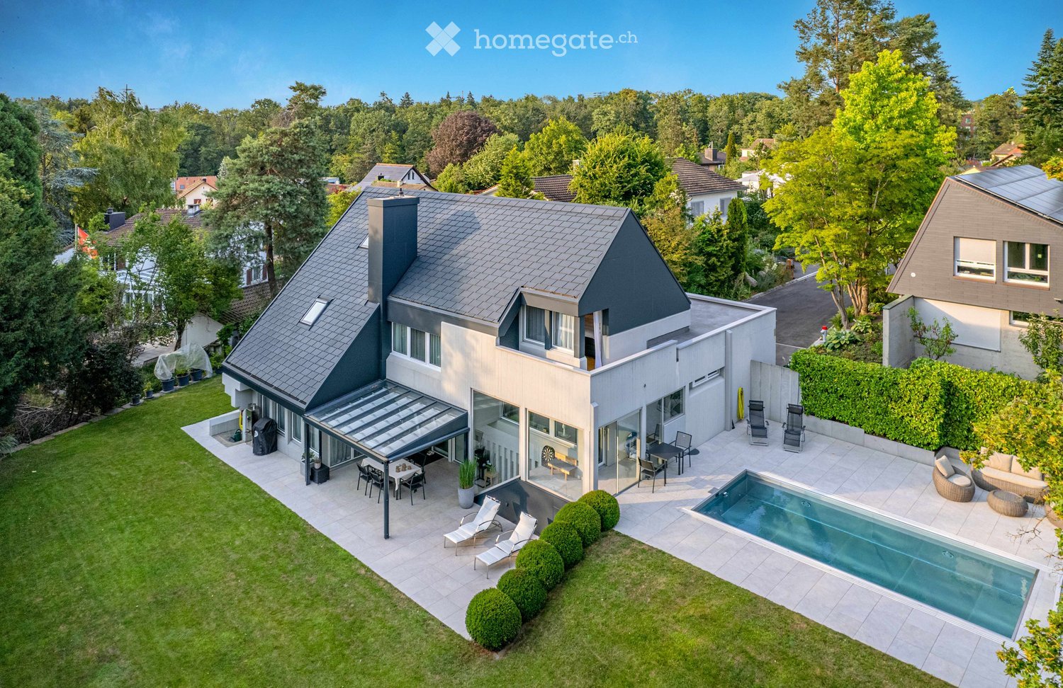 A modern two-story house with a grey roof and white walls, surrounded by a well-maintained garden, a swimming pool, outdoor seating, and a covered patio area.
