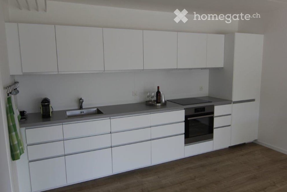 Modern kitchen with white cabinets, stainless steel appliances, and a grey countertop. Includes a sink, oven, refrigerator, and various small items like a coffee maker, wine glasses, and a bottle.
