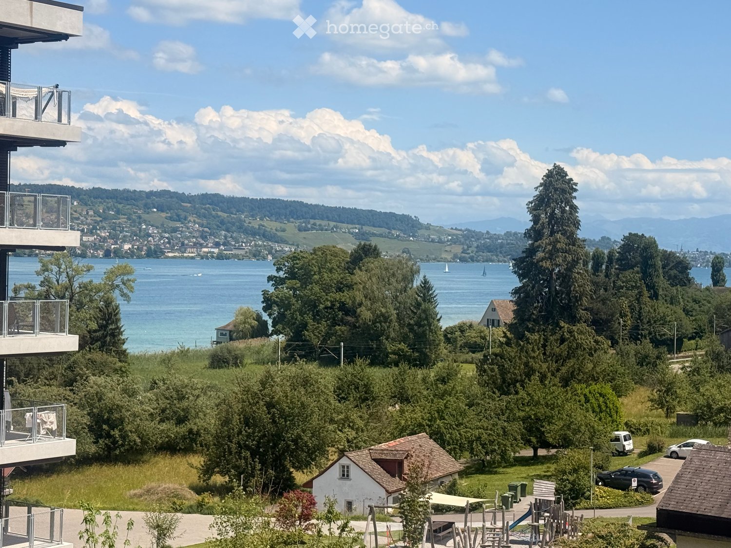 Balcony overlooking a scenic lake with sailboats, surrounded by trees and distant hills under a partly cloudy sky.
