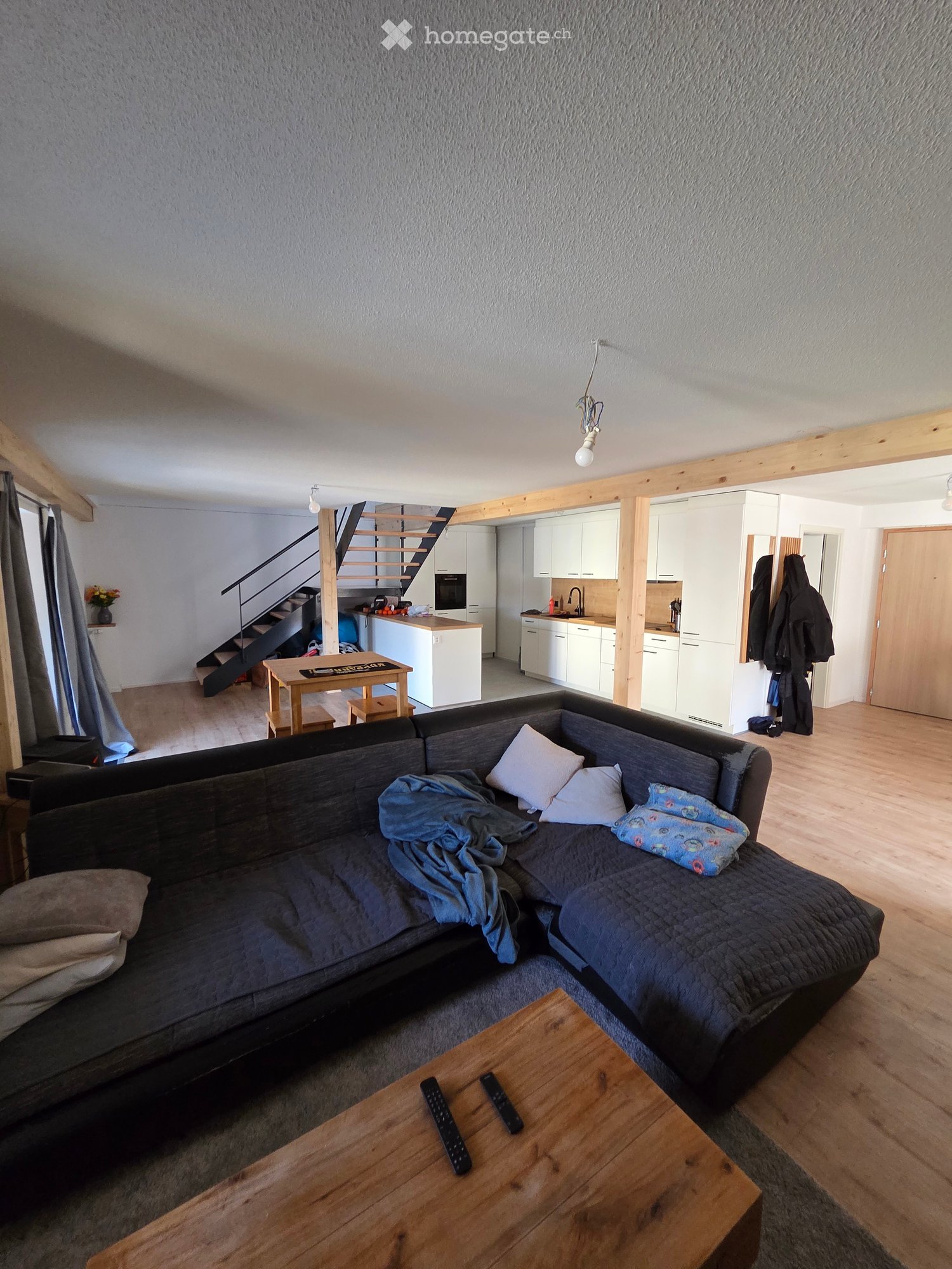 Living room with a grey sectional sofa, wooden coffee table, remote controls, pillows, and a blanket. The room features exposed wooden beams, a staircase leading to an upper level, and an open kitchen area with white cabinets, a sink, and an oven.