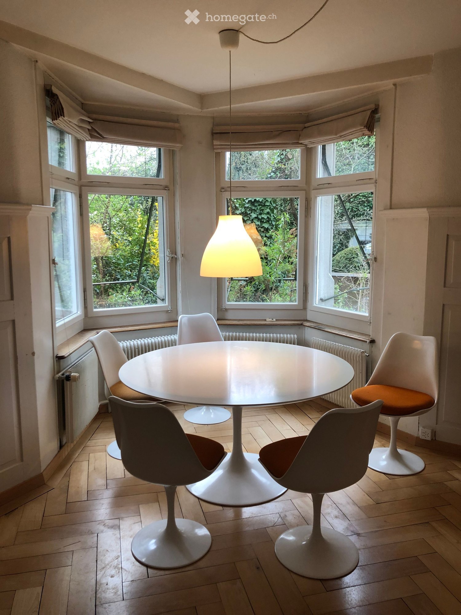 Bright room with large windows, wooden floor, round white table, four modern chairs, and a hanging lamp.