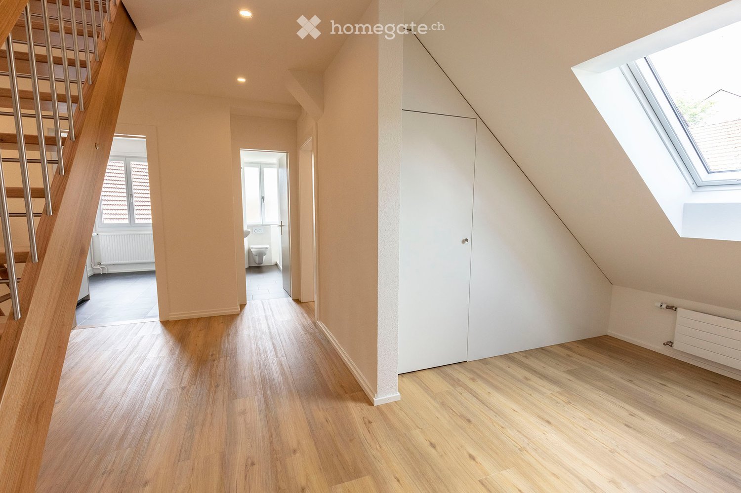 Bright, modern hallway with wooden flooring, white walls, and a staircase leading upwards. Features a built-in closet, a skylight window, and a view into a small bathroom with a toilet and sink.