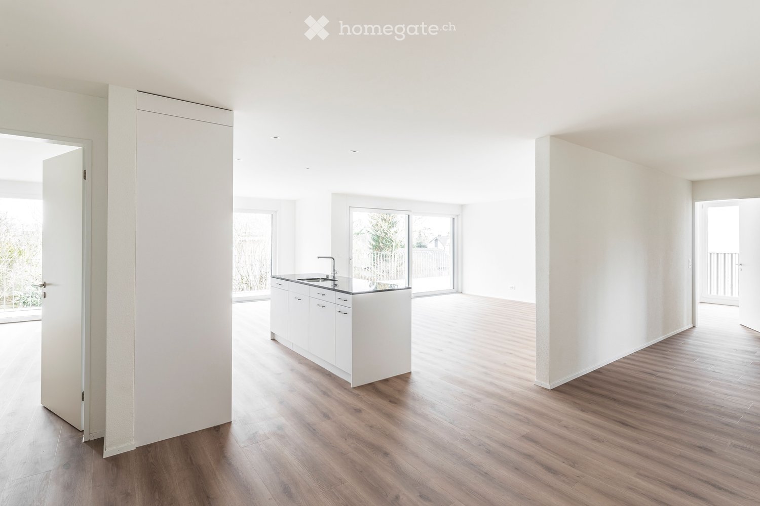 Open-plan unfurnished room with light-colored walls, wooden flooring, and a small white kitchen area with cabinets and a sink.