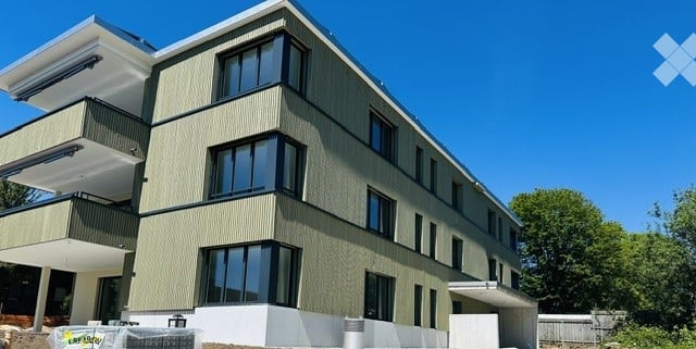 Modern multi-story building with large windows and balconies, surrounded by greenery.