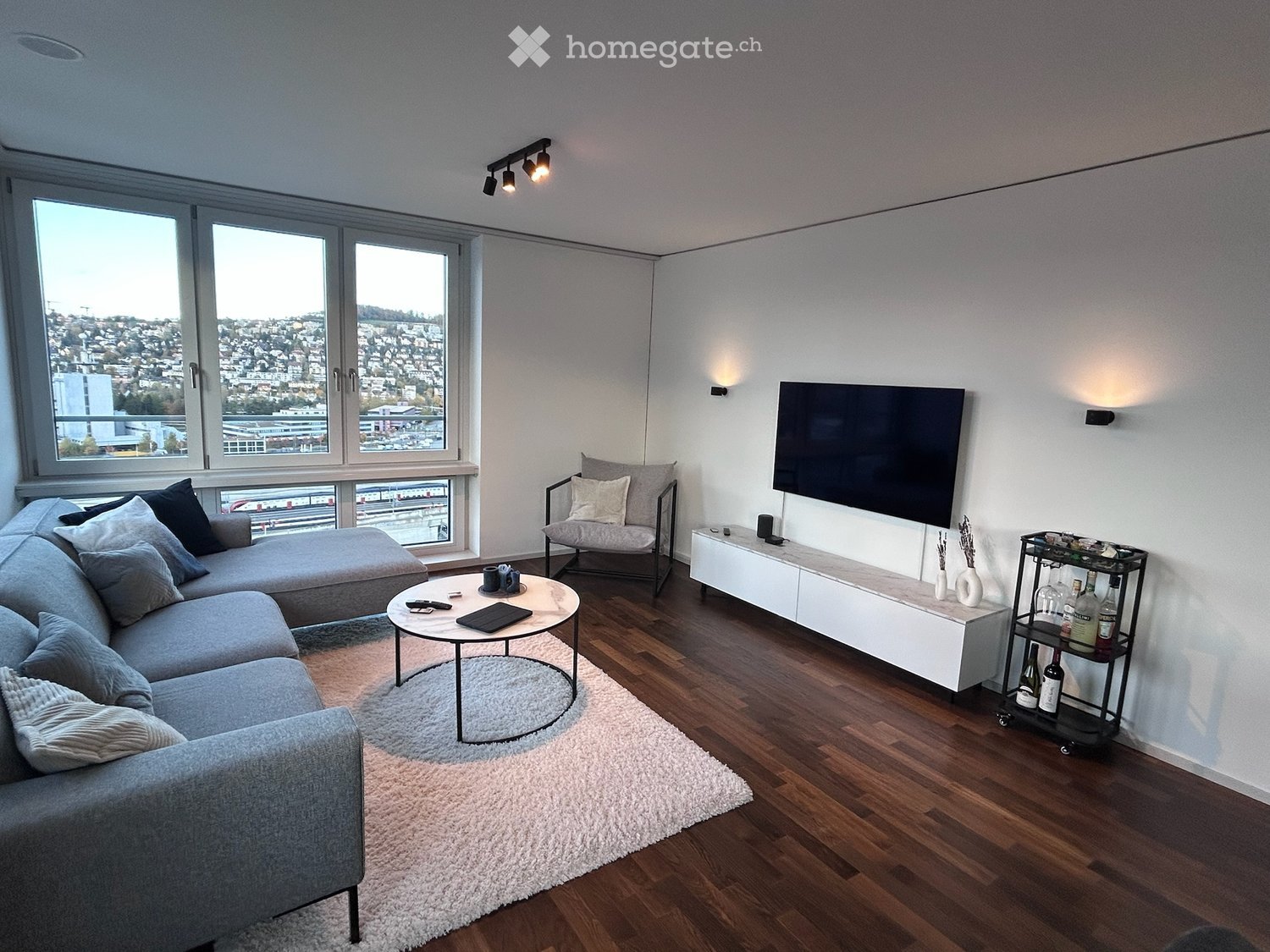 Modern living room with large windows offering a city view, grey sectional sofa, coffee table, armchair, TV, and a wine rack.