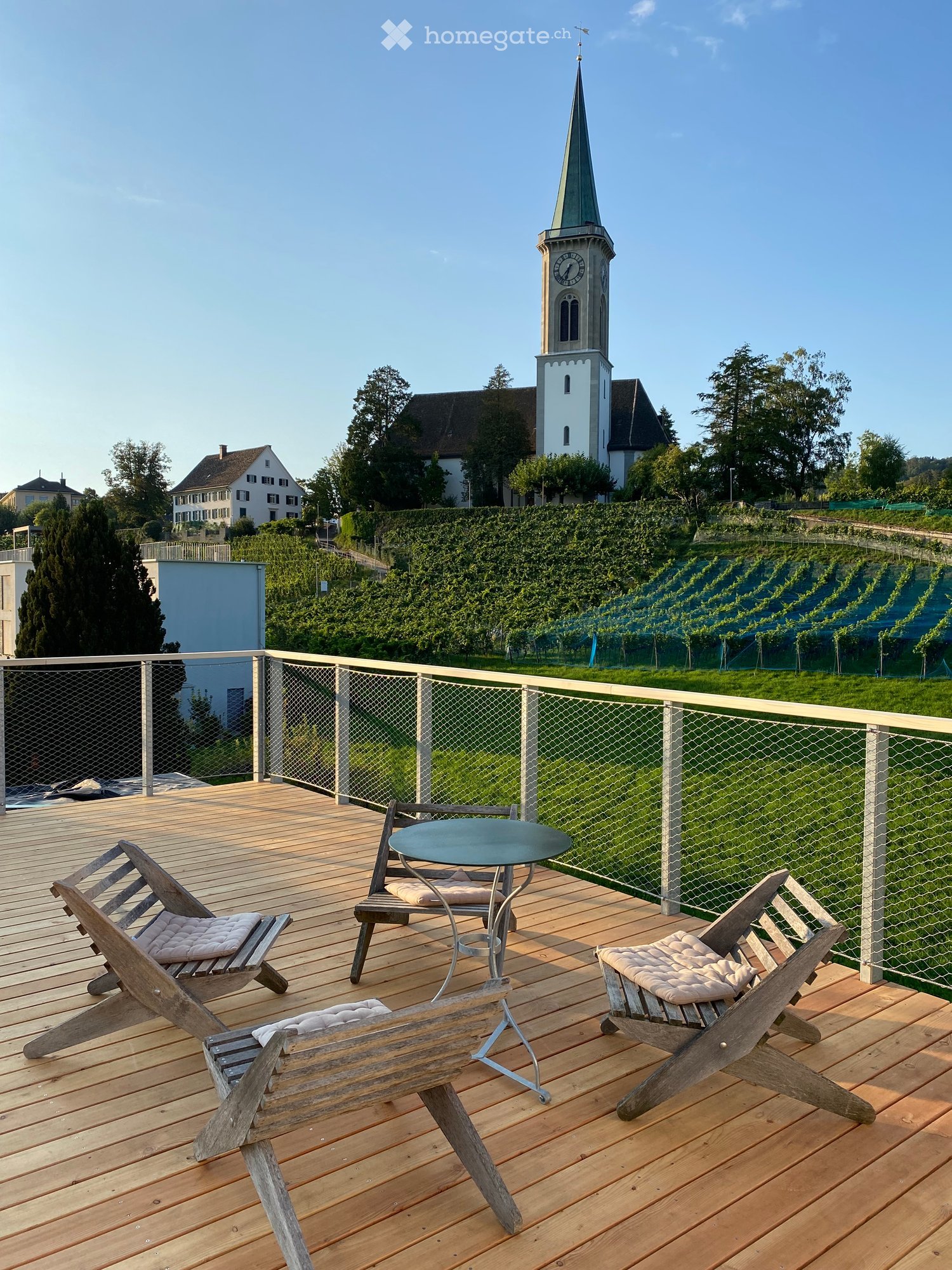 Wooden deck with a round table and two wooden chairs, overlooking a vineyard and a church tower.