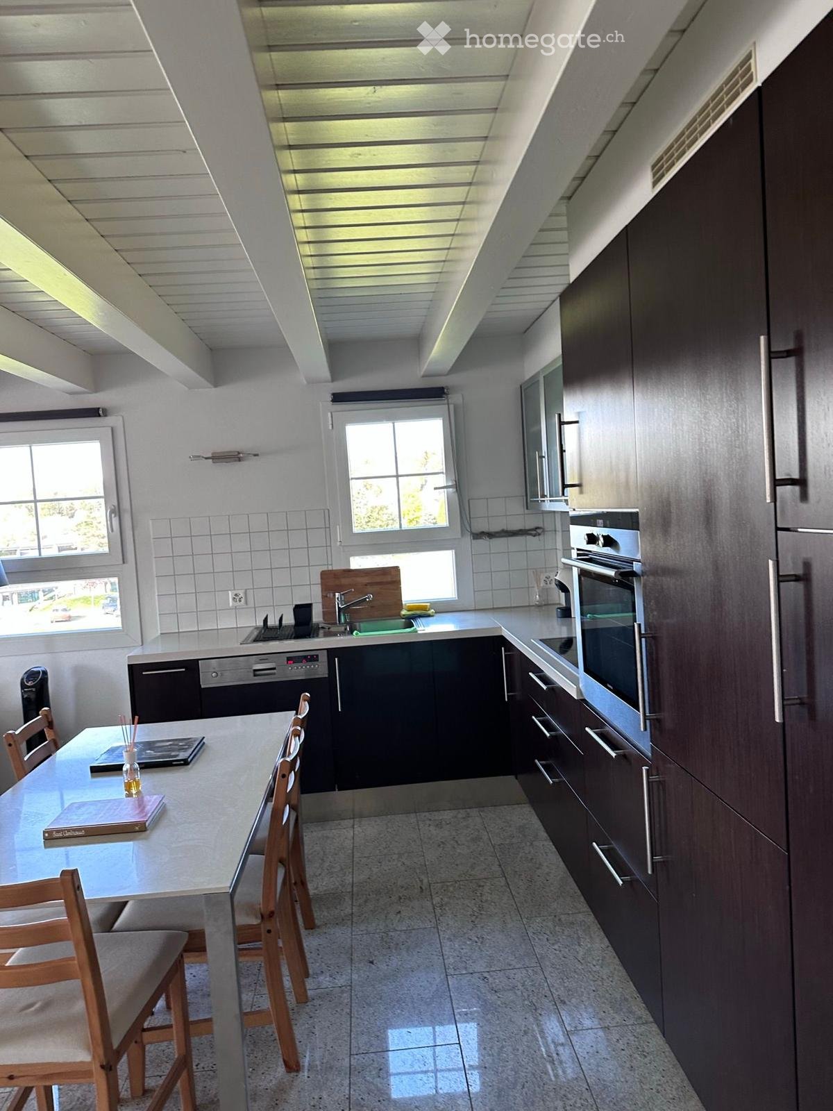 Modern kitchen with dark wooden cabinets, stainless steel appliances, a central island with a glass table and wooden chairs, and a sink with a faucet.