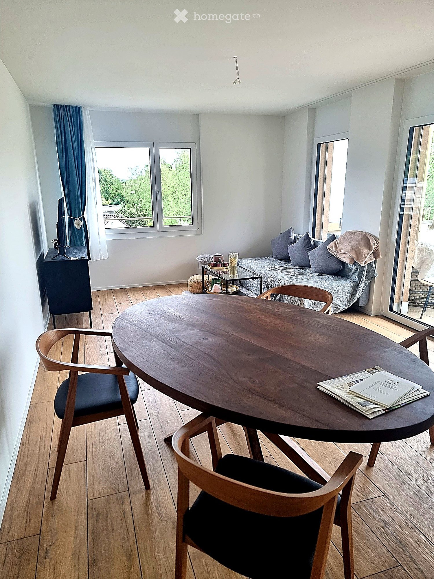 A modern, well-lit room with wooden flooring, a round wooden table with chairs, a sofa with pillows, a television on a stand, and a small side table with cups and a stool.