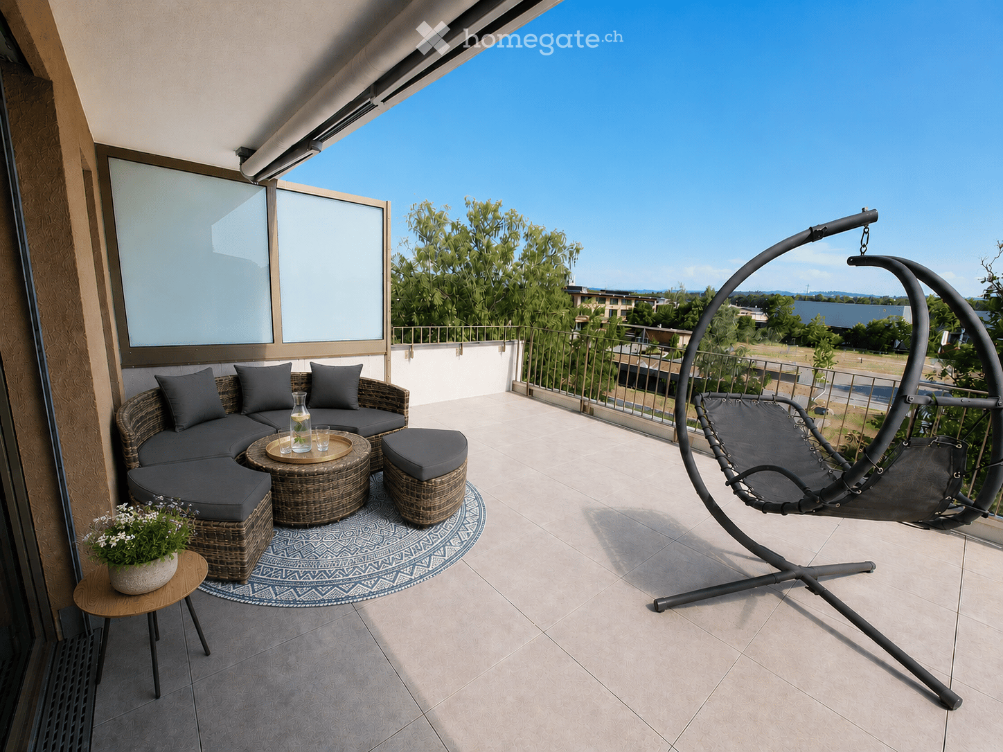 Balcony with a modern outdoor seating area, including a wicker sofa, round table, stools, and a hanging egg chair, overlooking a scenic view.
