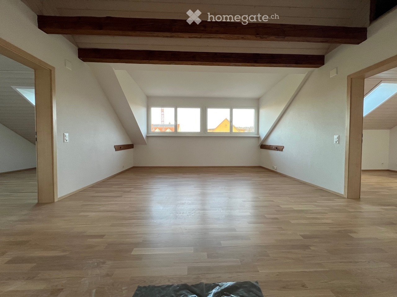 Empty attic room with wooden floors, exposed wooden beams, and large windows letting in natural light.