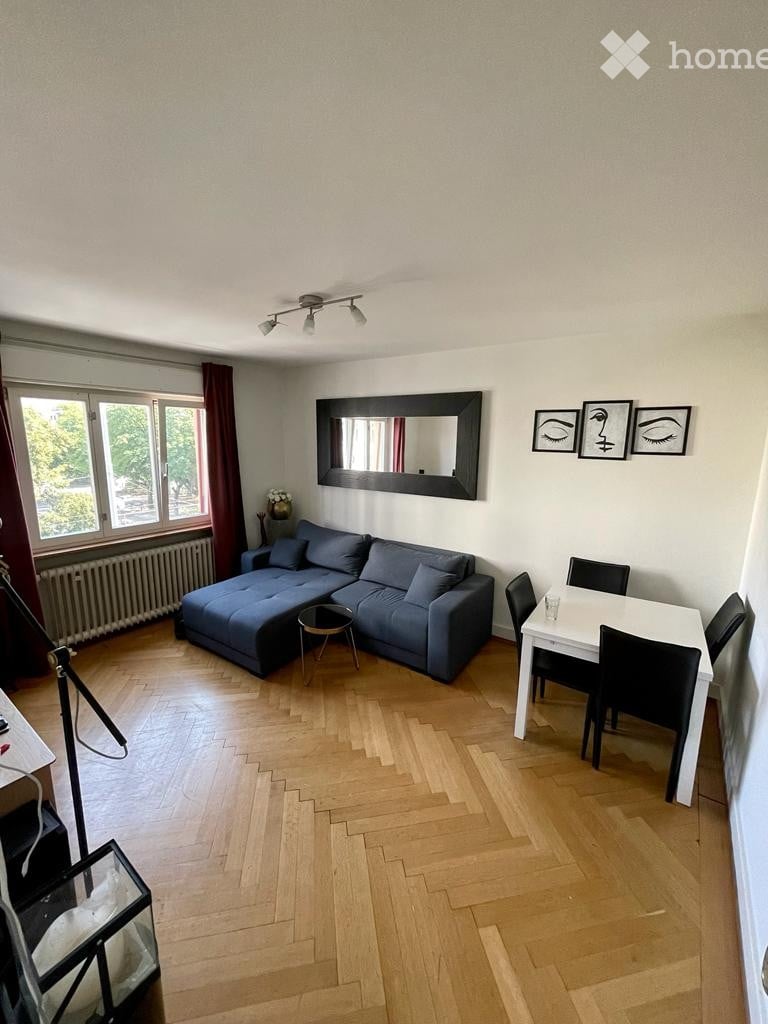 Modern living room with wooden flooring, large window, blue sectional sofa, small coffee table, dining table with chairs, and wall-mounted mirror.