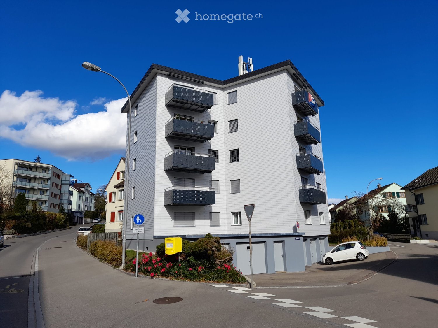 Modern multi-story apartment building with white facade, multiple balconies, and a small garden area at the base.