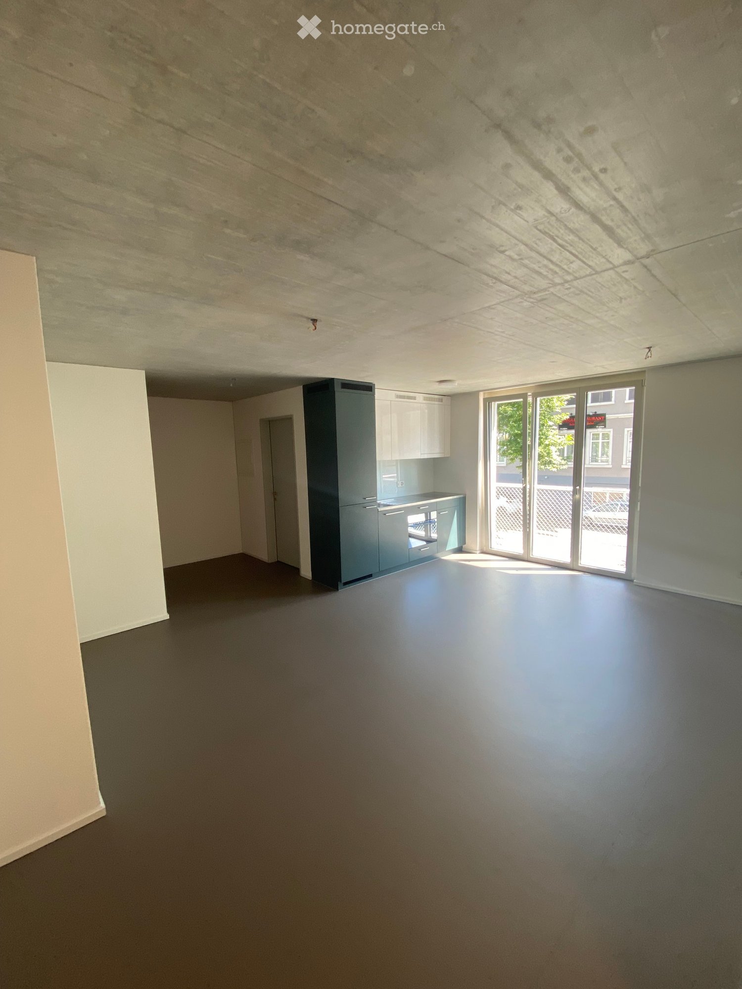 Empty room with grey carpeted floor, white walls, and a small kitchen area with cabinets and a countertop visible in the background. Large sliding glass doors lead to an outdoor area.