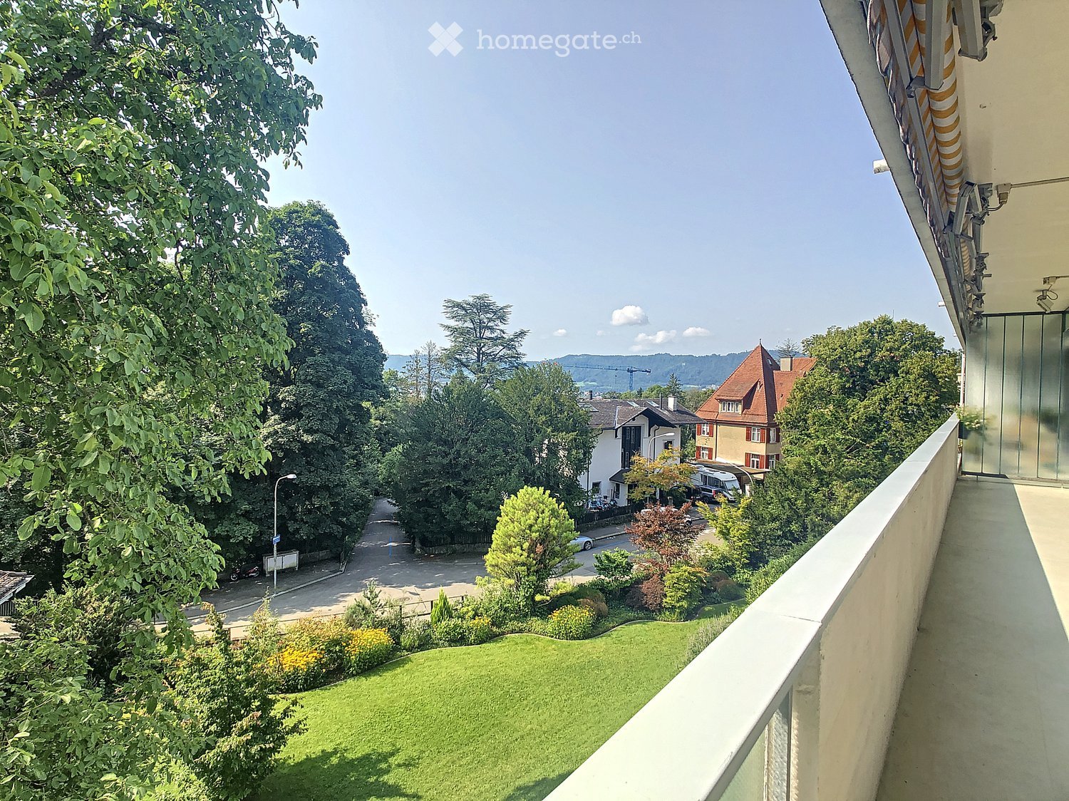 Balcony overlooking a green lawn, trees, and a street with houses and parked cars in the background.