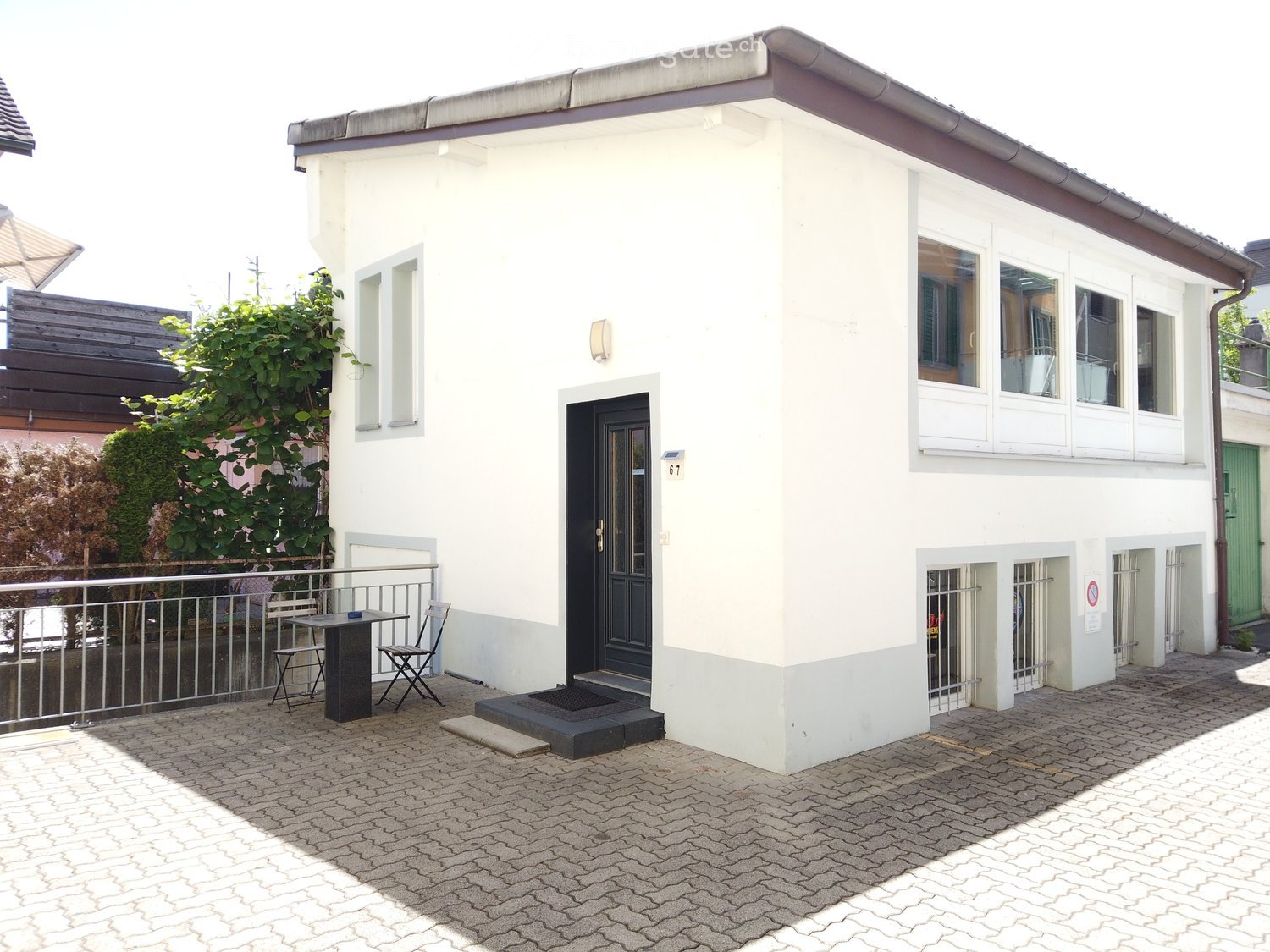 Two-story white building with a small outdoor seating area featuring a table and chairs, a metal railing, and a paved cobblestone area.