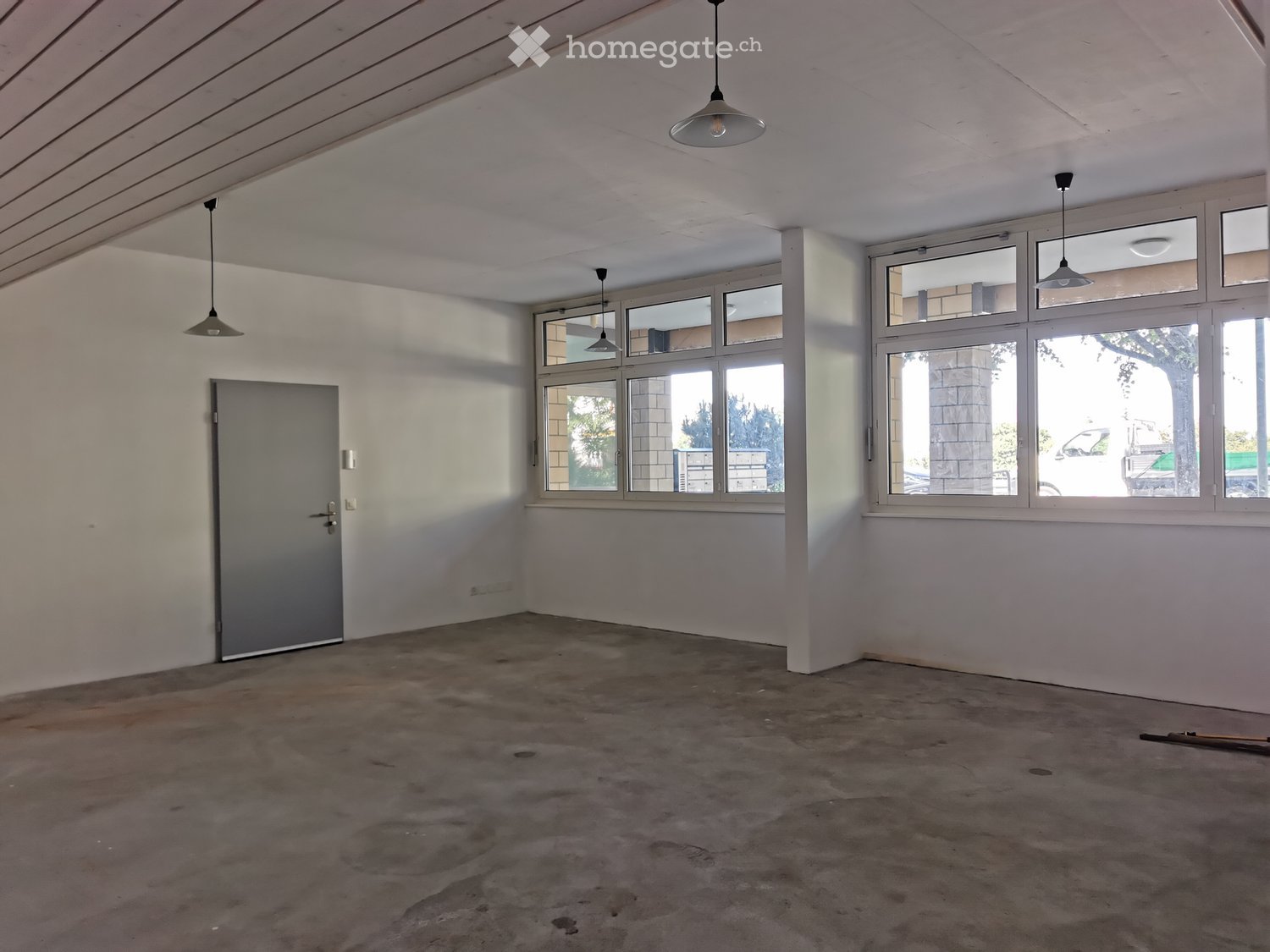 Empty room with concrete floor, white walls, large windows, and pendant lights.