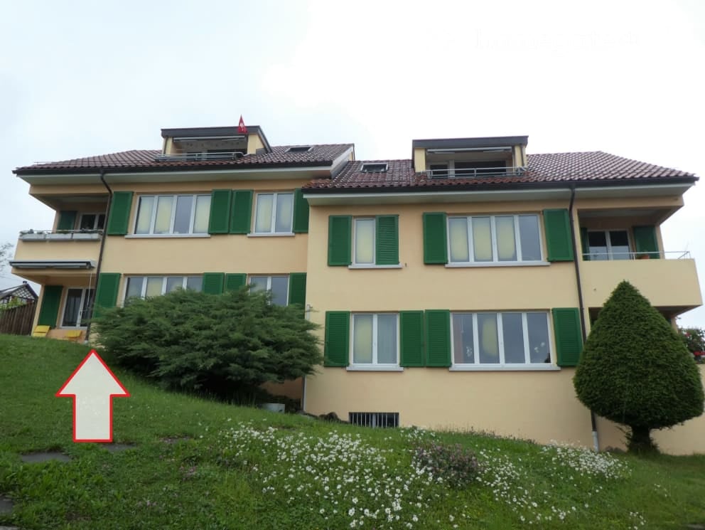 House. Two-story house with yellow walls, green shutters, and multiple windows. The house is situated on a raised ground floor with a well-maintained garden in front.
