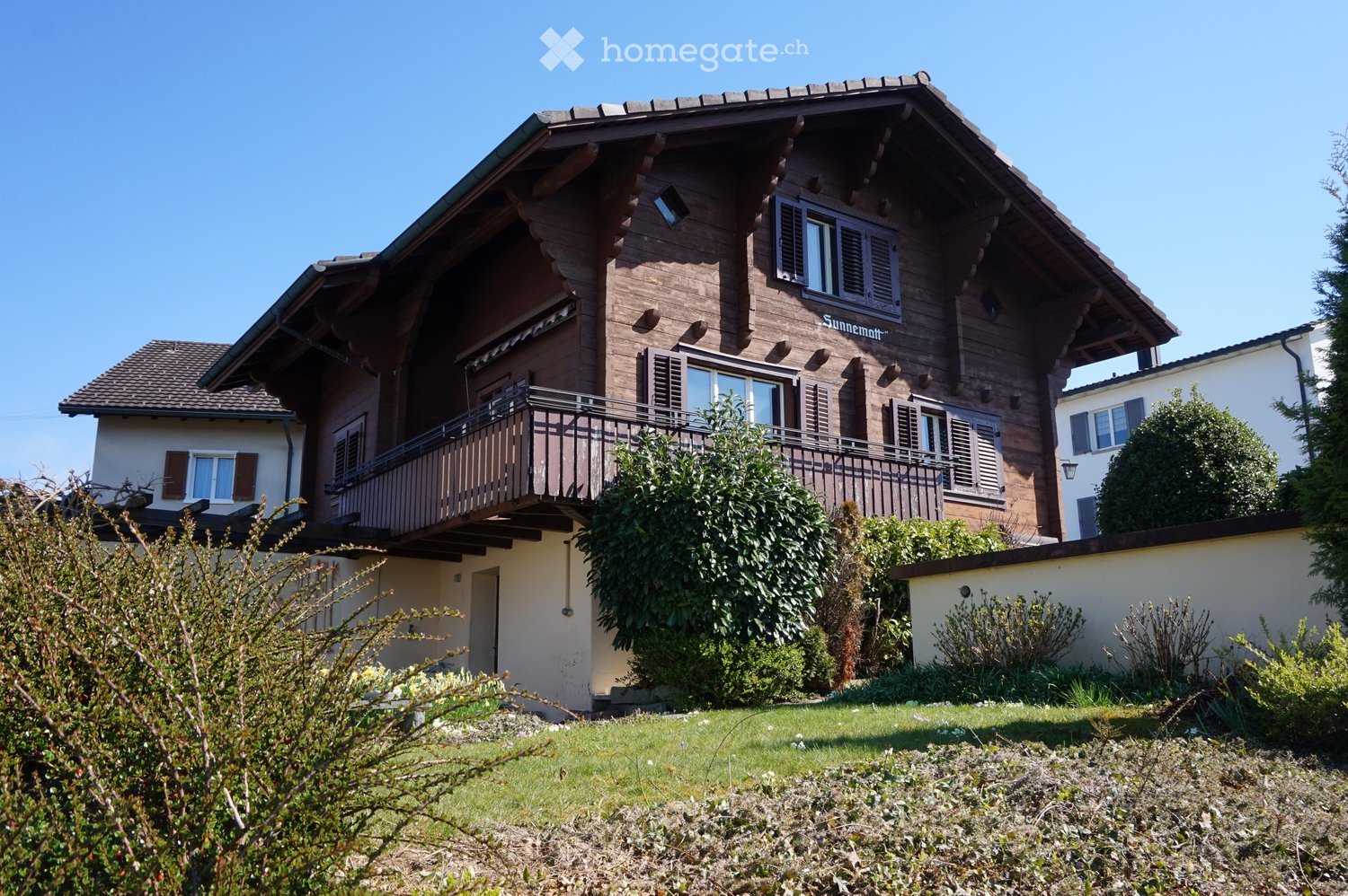 A two-story wooden house with a steeply pitched roof, multiple windows with shutters, and a balcony on the upper level. The house is surrounded by a well-maintained garden with various shrubs and trees.