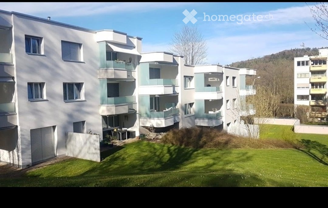 Modern multi-story residential building with white and light green facade, multiple balconies, and a well-maintained lawn in front.