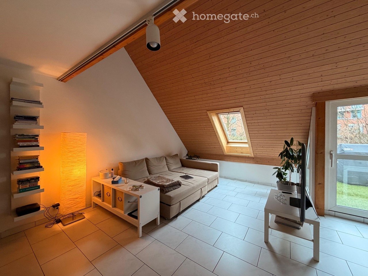 Modern living room with a slanted wooden ceiling, white tiled floor, and large windows. Features a grey sectional sofa, coffee table, side table, floor lamp, bookshelf, potted plant, and television.