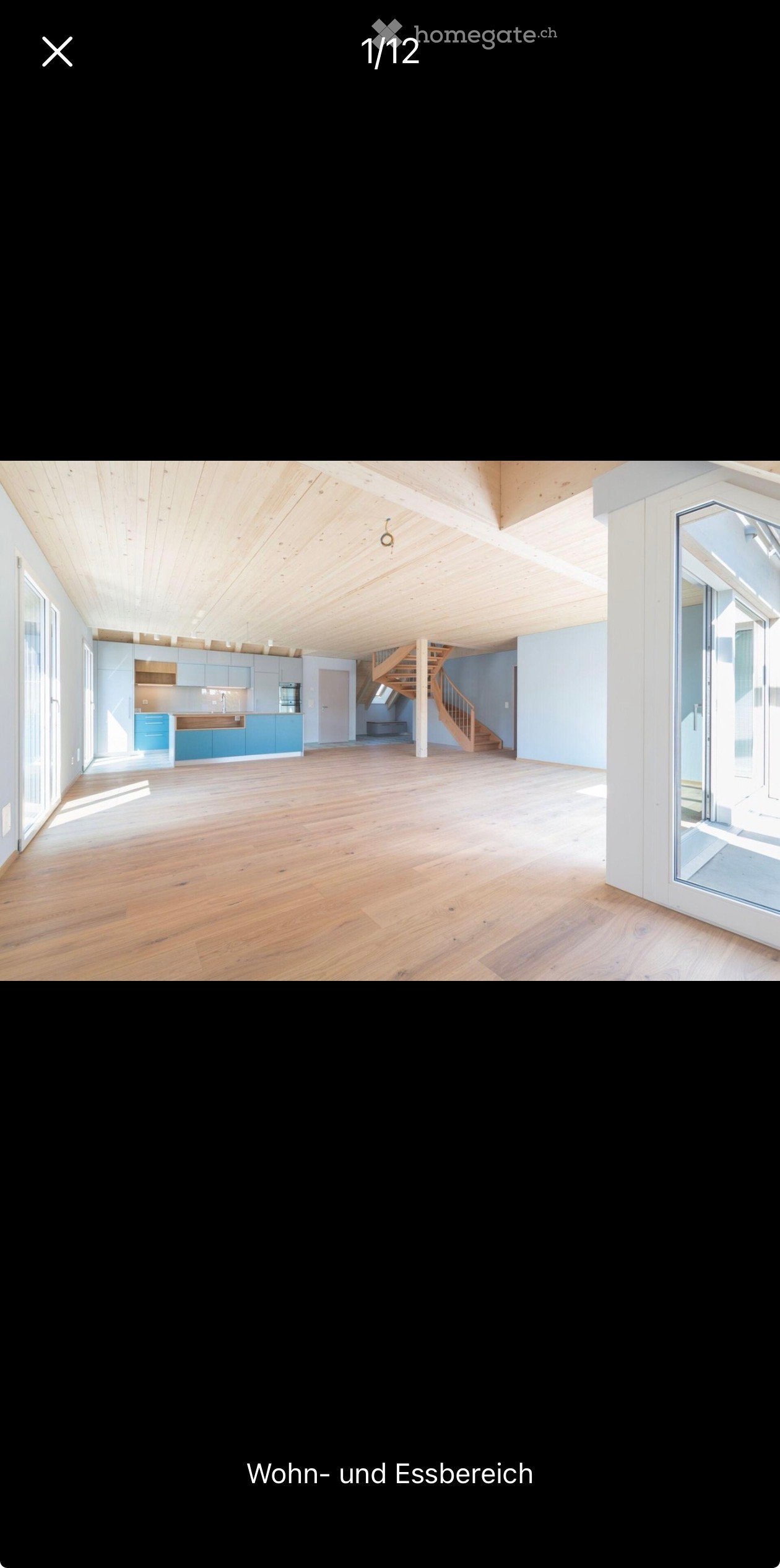 Open-plan living and dining area with light wooden flooring, exposed wooden ceiling beams, and modern kitchen cabinets. A wooden staircase leads to an upper level.