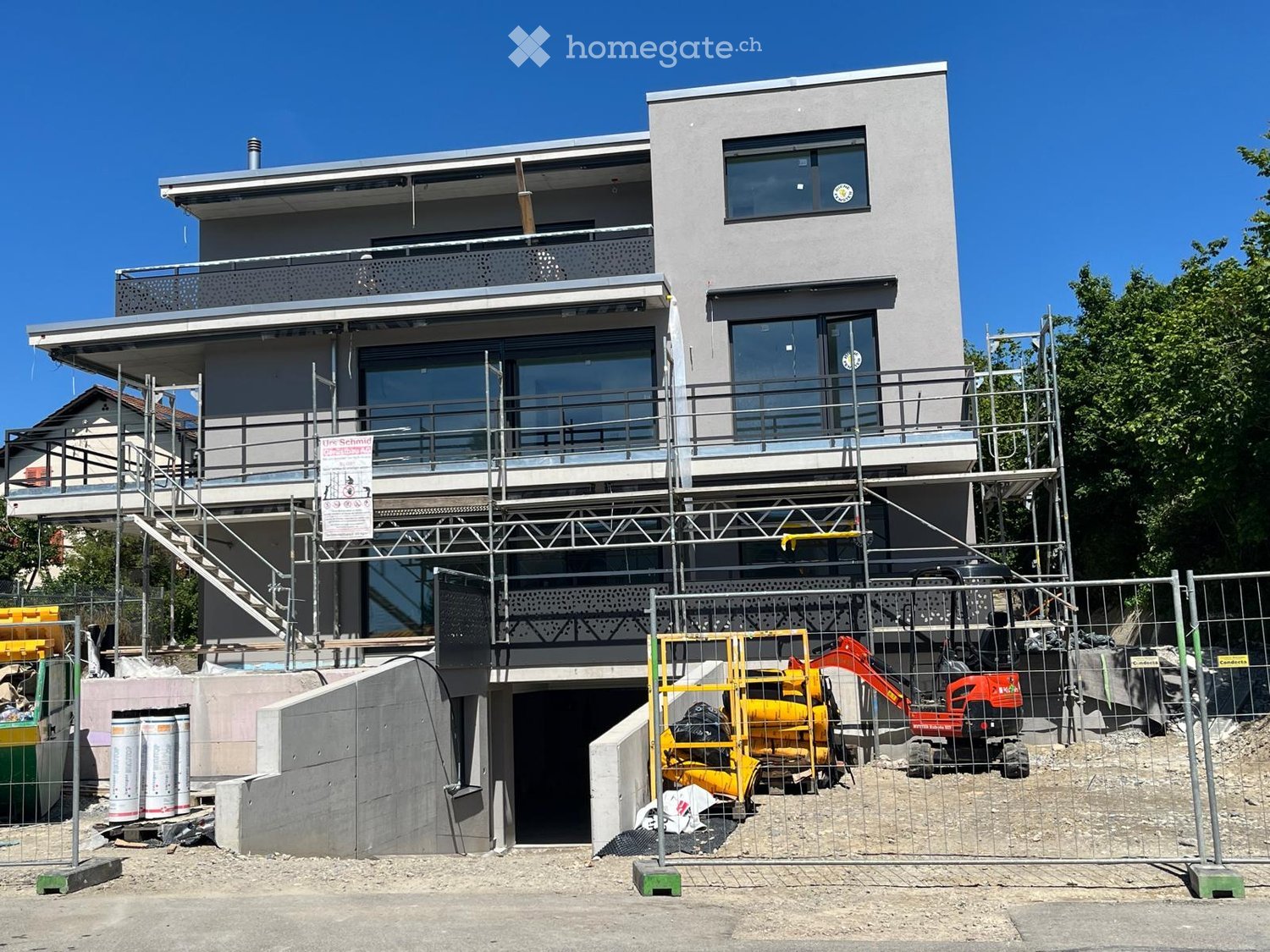 Modern two-story house under construction with scaffolding, large windows, and a balcony area. Construction equipment and materials are visible in the foreground.