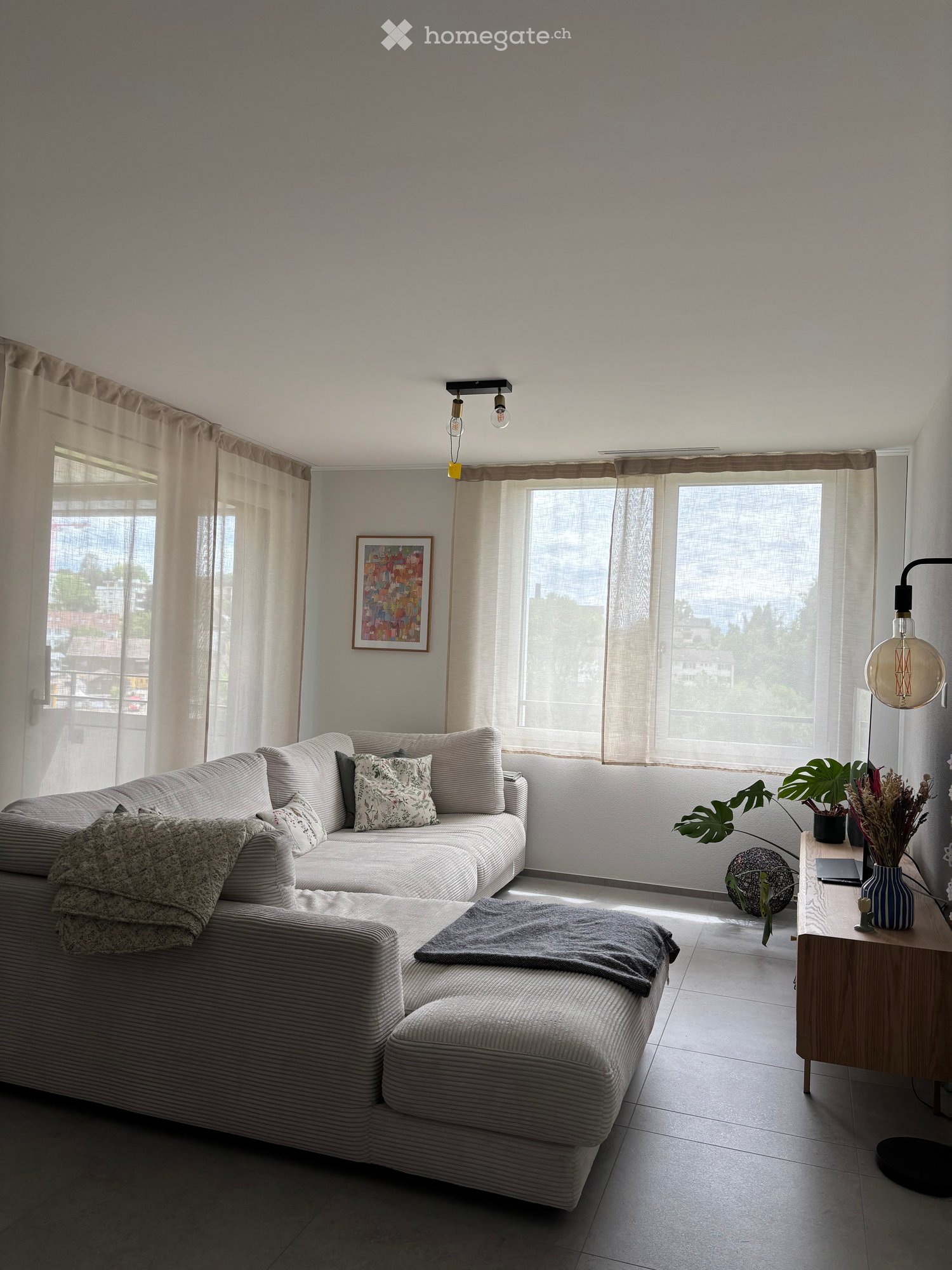 Modern living room with a grey sectional sofa, coffee table, side table with plants and a laptop, and large windows with sheer curtains.